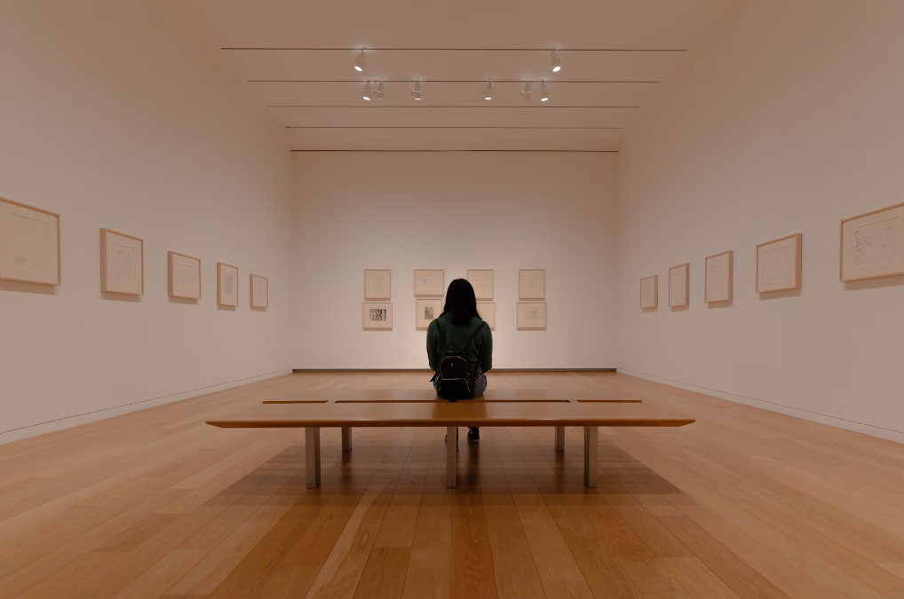 a gallery visitor wearing a backpack sitting down in a white room with several prints along the wall